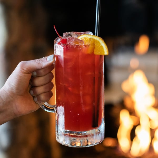 Hand holding a red cocktail with orange slice and cherry, with a fireplace in the background.