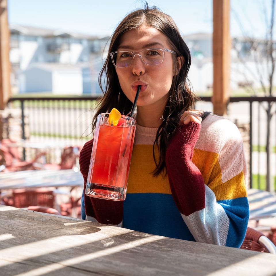 A woman in a colorful sweater sips a red drink with a straw on a sunny patio.