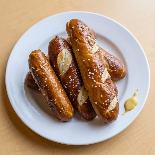 A white plate holds several golden-brown pretzel sticks, sprinkled with coarse salt, and a dollop of mustard.
