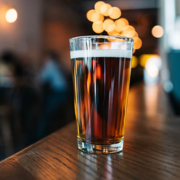 A glass of amber beer with a foamy head sits on a wooden bar, with bokeh lights in the background.