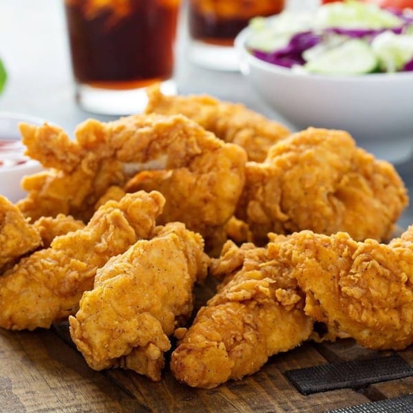 Crispy fried chicken tenders on a wooden board, with drinks and salad in the background.